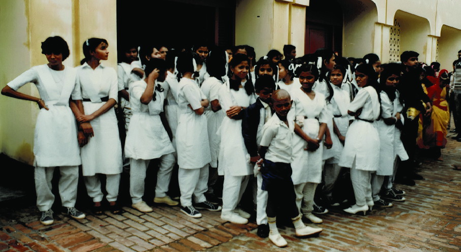 A long line of children in white uniform standing along a verandah