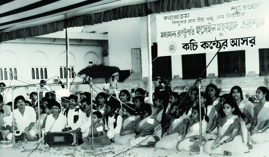 Children seated on the ground before a stage of microphones, under a banner reading “কচি কণ্ঠের আসর”