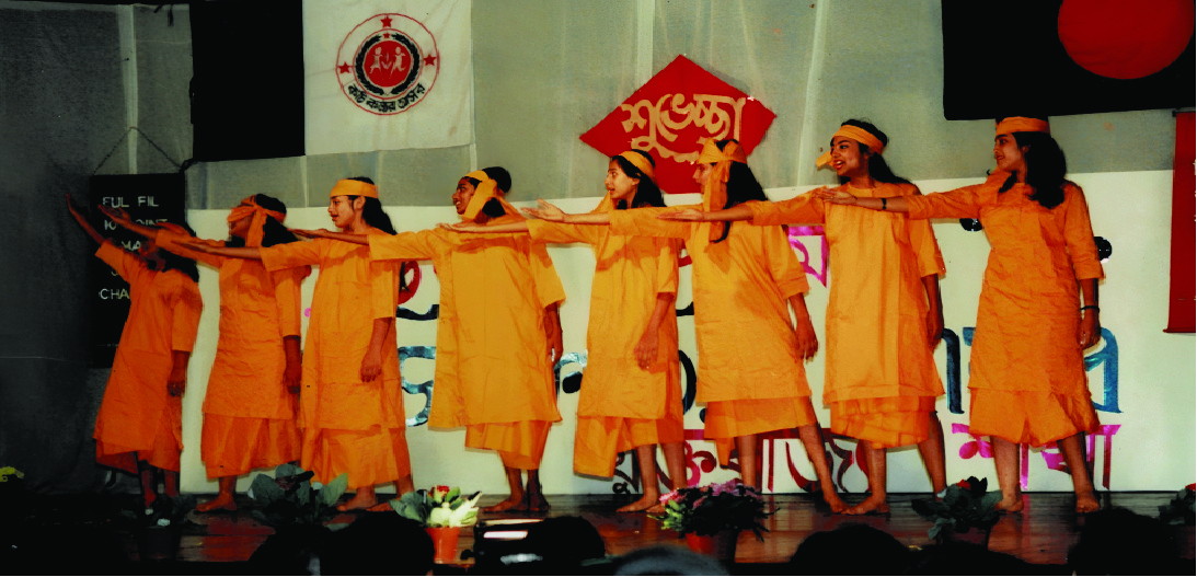 A line of children in orange costume dancing on a stage with their arms linked