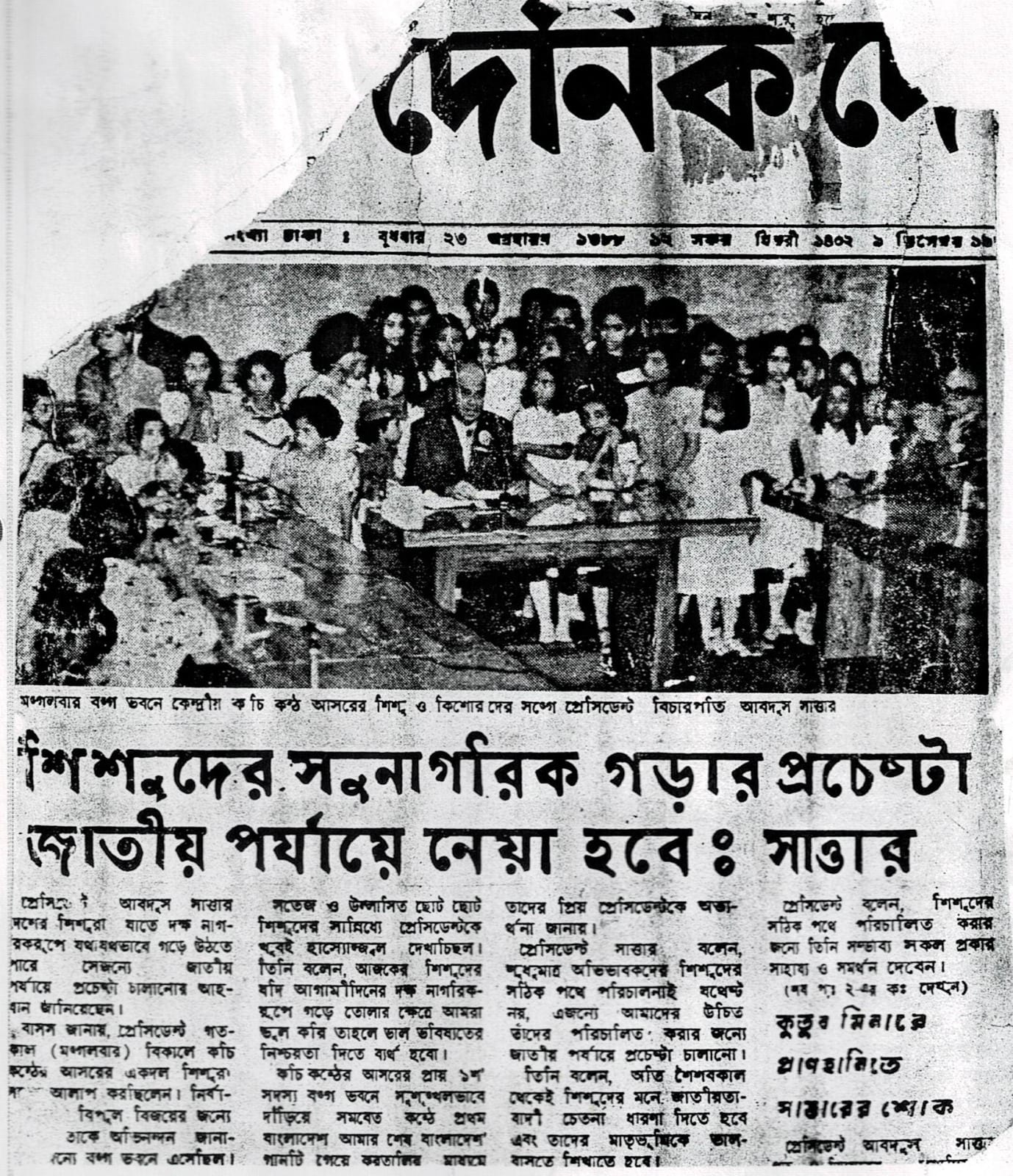 A newspaper front page: a man seated at a desk with children standing several rows deep on both sides of him
