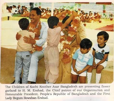 A colour photograph: small children placing a flower garland over a seated man, with a woman in a sari and more children beside them