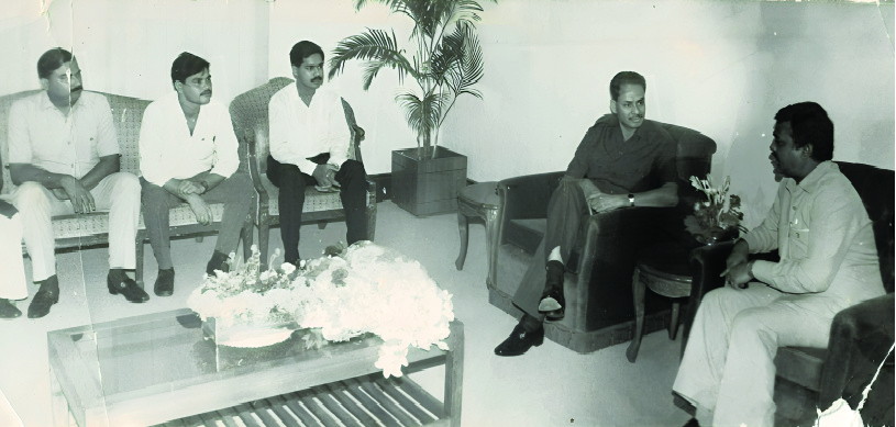 Men seated in a reception room, one offering flowers across a low table