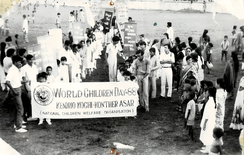 A procession crossing a field behind a banner reading “WORLD CHILDREN DAY-88, KENDRIO KOCHI-KONTHER ASAR (NATIONAL CHILDREN WELFARE ORGANISATION)”, with children carrying placards behind it