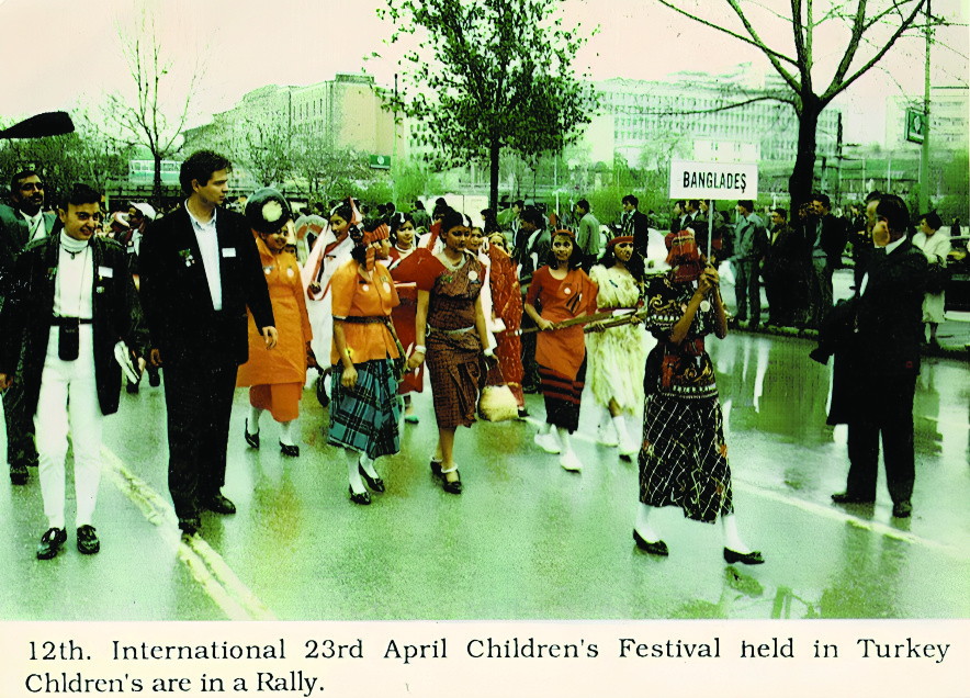 A rally of children and adults in national dress walking behind a placard reading BANGLADESH, under the printed caption “12th. International 23rd April Children’s Festival held in Turkey”