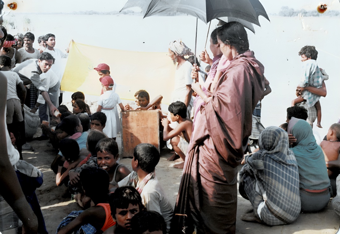Families gathered by a river for a relief distribution, a man handing out supplies from a sack