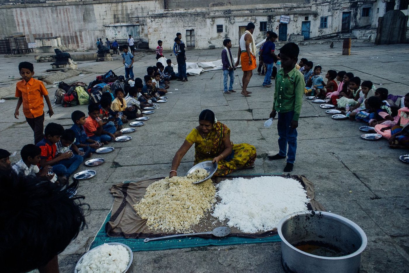 Rice being served from large pots to rows of children seated on the ground