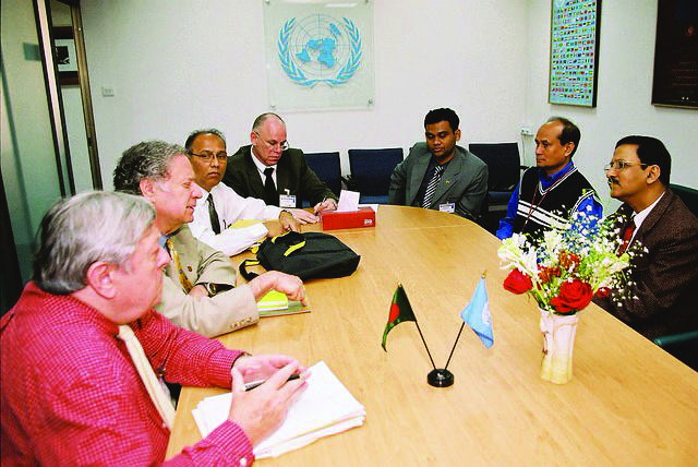 Delegates seated around a conference table beneath the United Nations emblem, with the flag of Bangladesh standing on the table